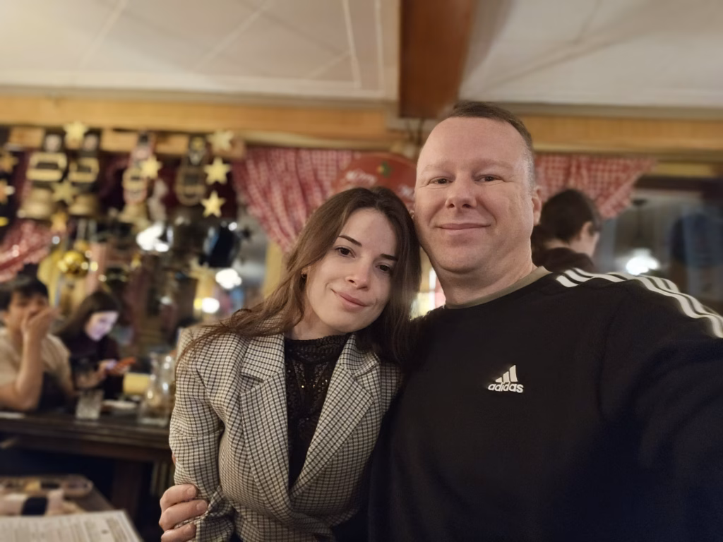 Ryan and Samira taking a selfie at a restaurant table with a red-checked cloth and a souvenir display behind, on the arrival evening
