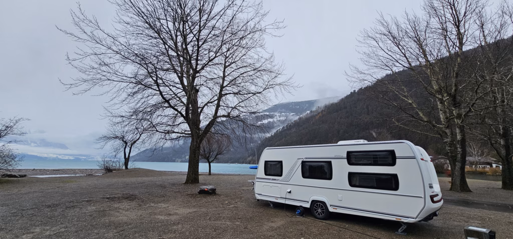 A white touring trailer pitched on the bare pebbly shore of Lake Thun with bare winter trees and cloud-wrapped alps behind