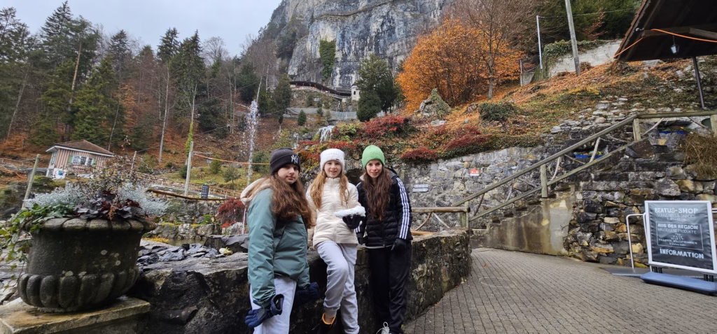 Three girls in winter jackets standing on the path at the foot of the St. Beatus cliff with the cave entrance and autumn foliage above
