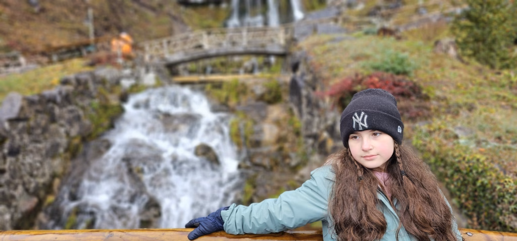 Lulu in a dark beanie and green jacket leaning on a wooden railing with the St. Beatus waterfall and a stone footbridge behind