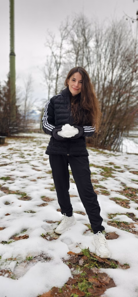 Samira standing on snowy ground holding a snowball and smiling, with bare trees behind at a winter rest stop
