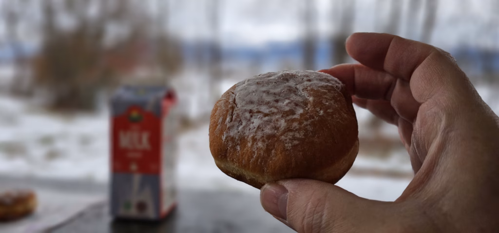 A hand holding a powdered-sugar Berliner doughnut with a carton of milk and a snowy lakeside behind