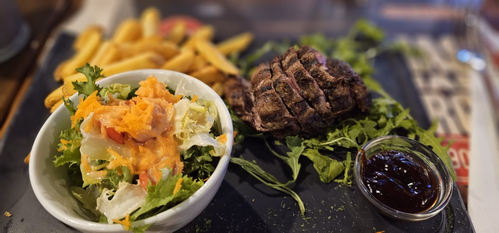 Sliced grilled steak with fries, a side salad in a white bowl and a ramekin of sauce on a slate plate at a restaurant