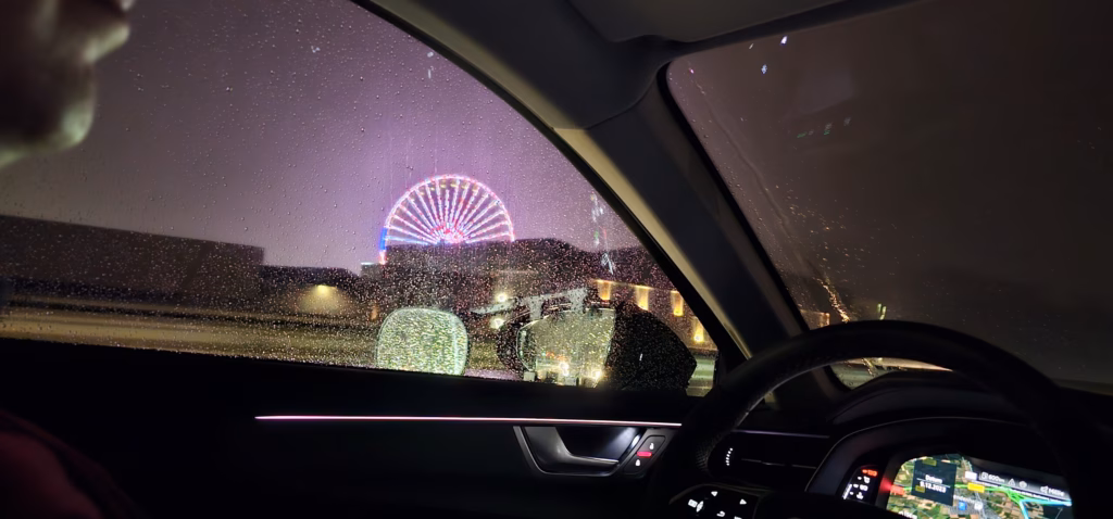 A brightly lit Ferris wheel seen at night through a rain-spotted car window with the steering wheel and navigation screen inside the car