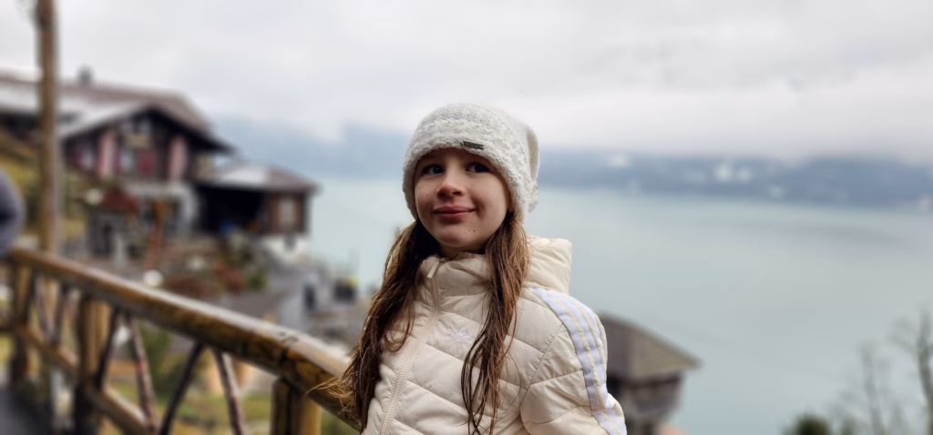 Sahryah in a white knit beanie and cream jacket at a wooden railing with Lake Thun behind