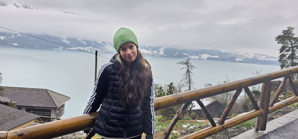 Samira in a green beanie and black jacket at a wooden railing with the turquoise Lake Thun and snowy far shore behind