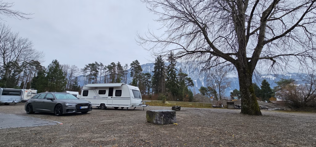 A grey Audi and white Fendt caravan parked on a gravel Stellplatz under a large bare tree with snowy ridges behind