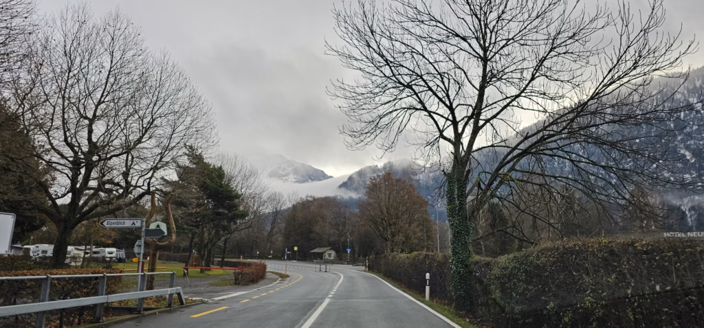 A road curving past the campground with parked caravans and bare trees and cloud-wrapped alps behind