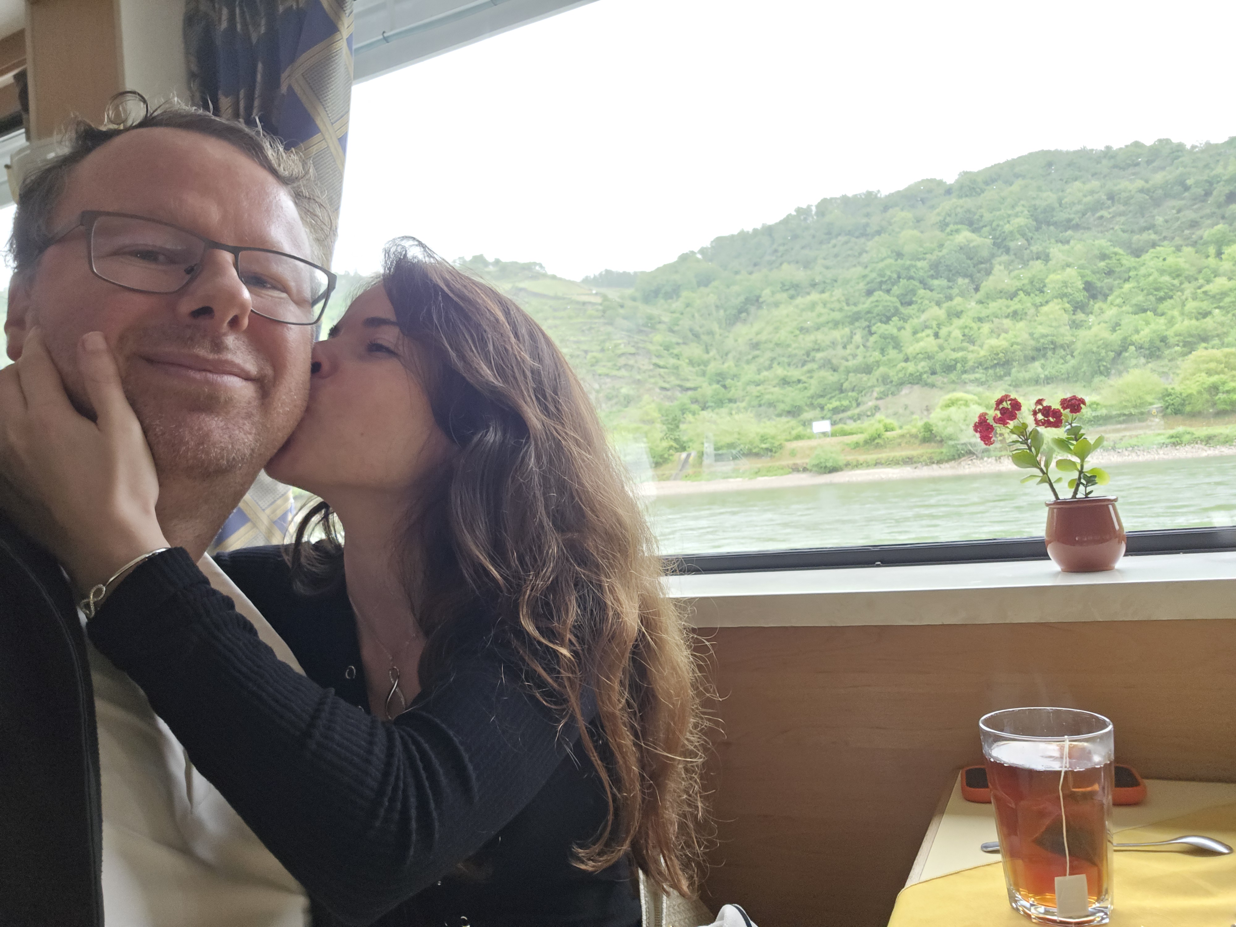 Ryan and Samira sharing an anniversary kiss in the KD saloon, Rhine gorge visible through the window