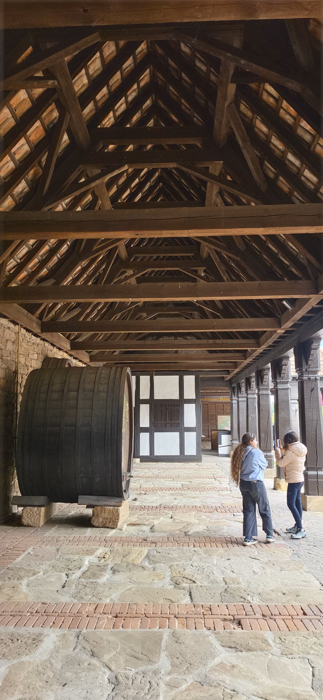 Wooden roof beams of the Vinothek loft at the Deutsches Weintor with Samira and Lulu standing next to a giant Fuder barrel