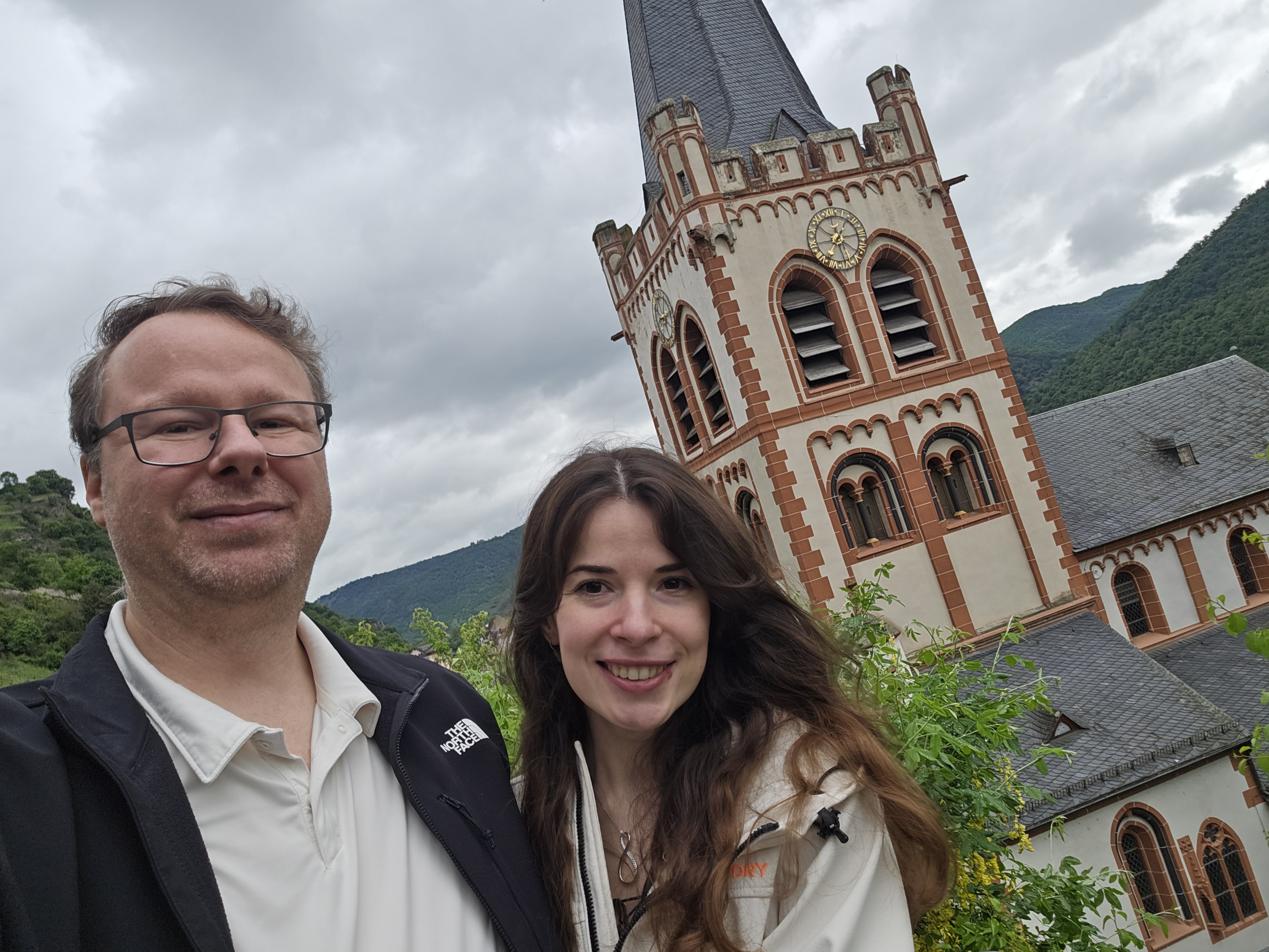 Ryan and Samira selfie above the Bacharach rooftops, St. Peter Kirche tower behind, Burg Stahleck visible on the ridge above, anniversary morning
