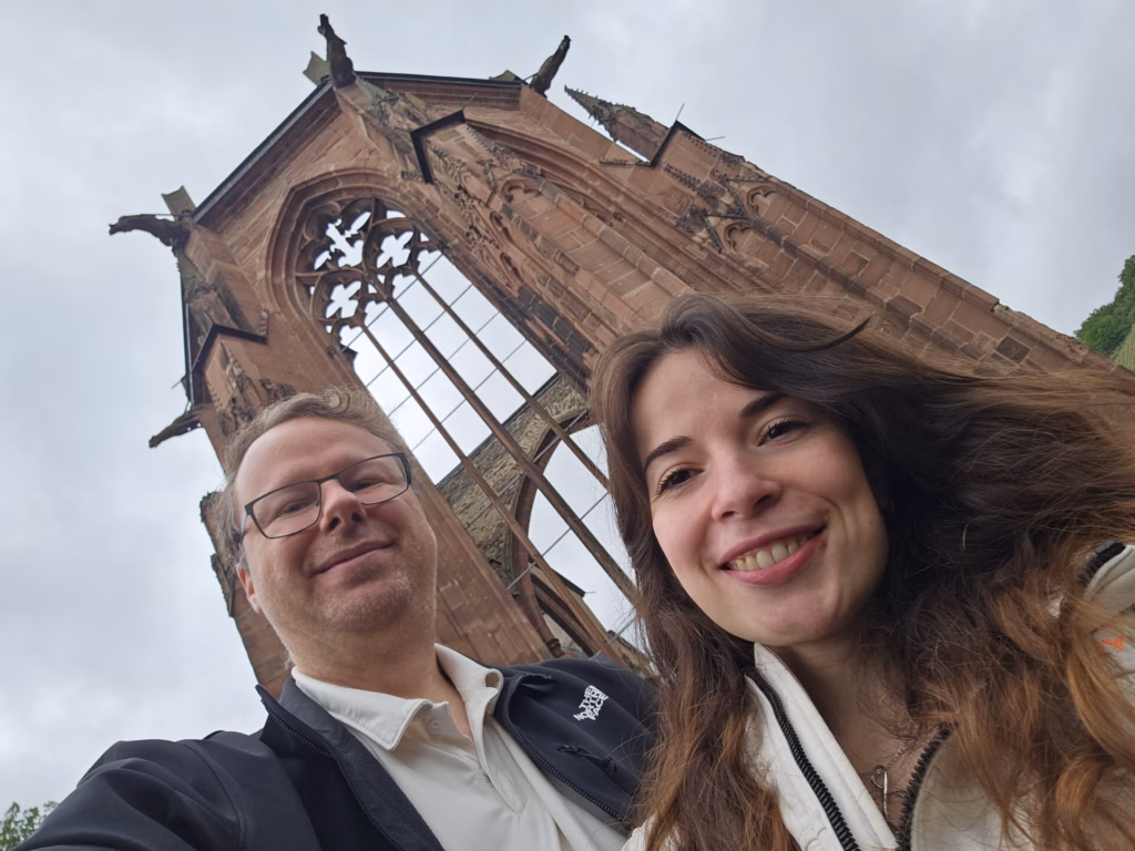 Ryan and Samira taking a selfie below the roofless red-sandstone Gothic ruin of the Wernerkapelle above Bacharach
