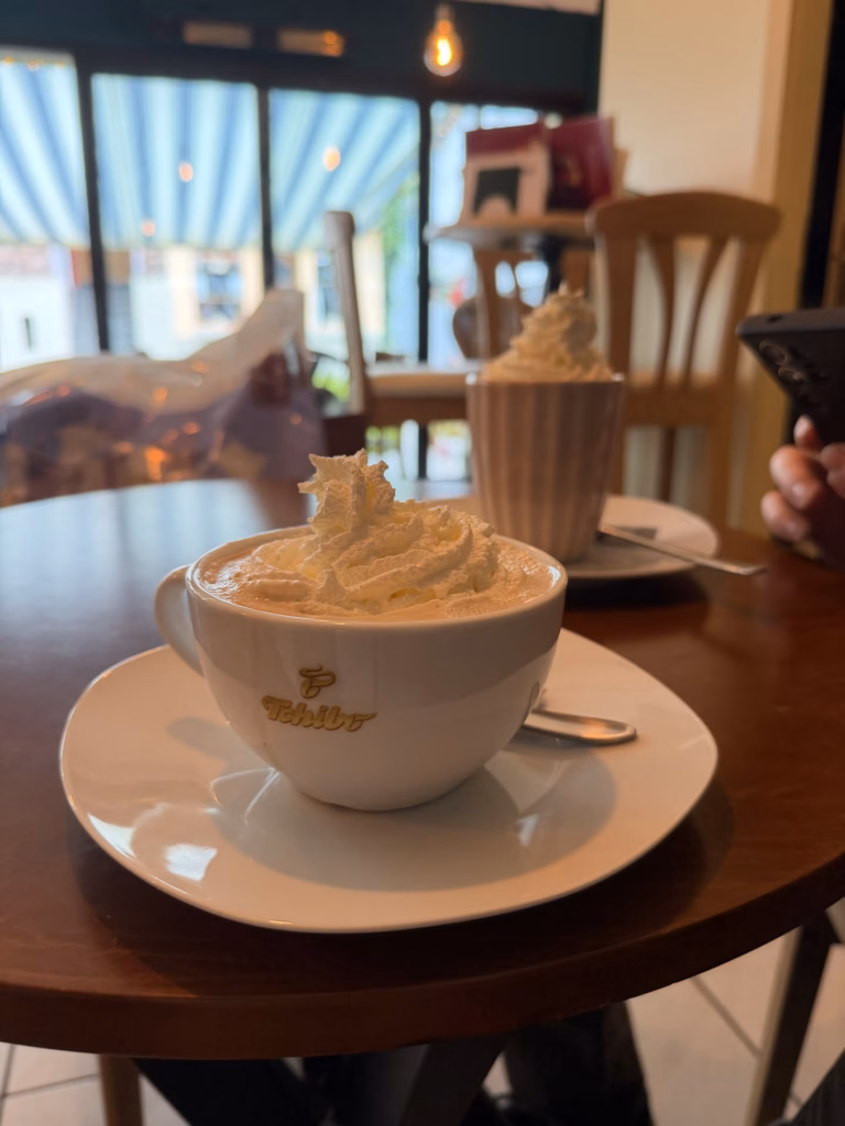 A cup of coffee topped with a tall swirl of whipped cream on a Tchibo saucer at a cafe table, a second cream-topped drink behind