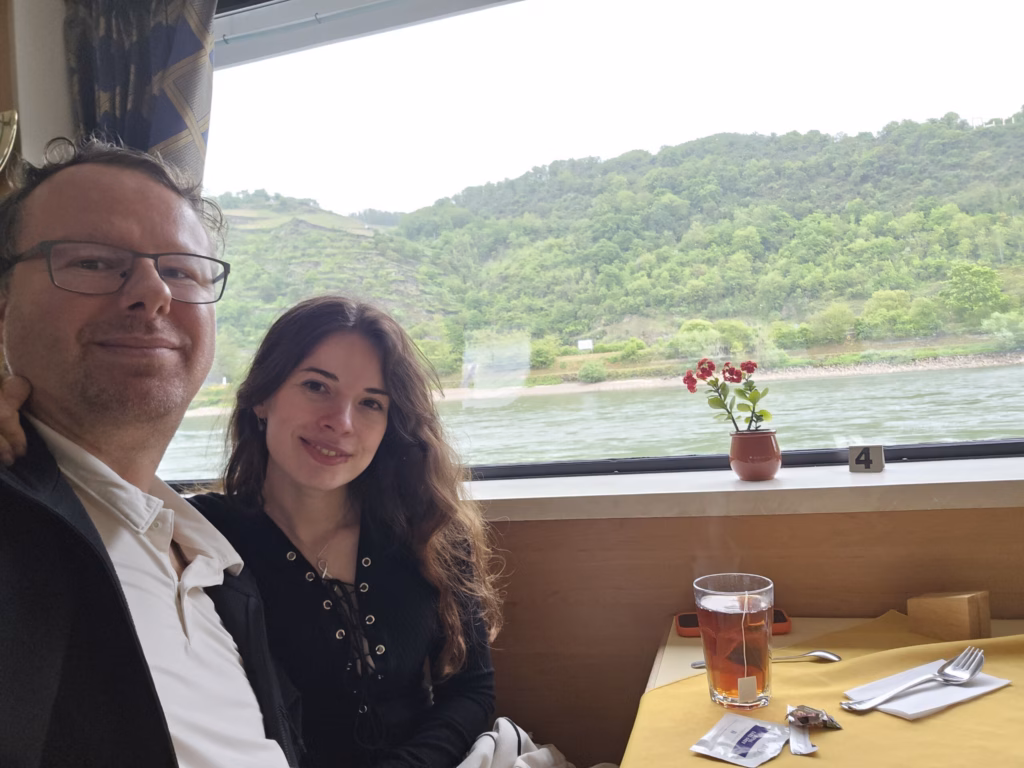 Ryan and Samira at a window table on the covered deck of the KD cruise boat, a glass of tea and a small red geranium on the sill, green Rhine bank beyond