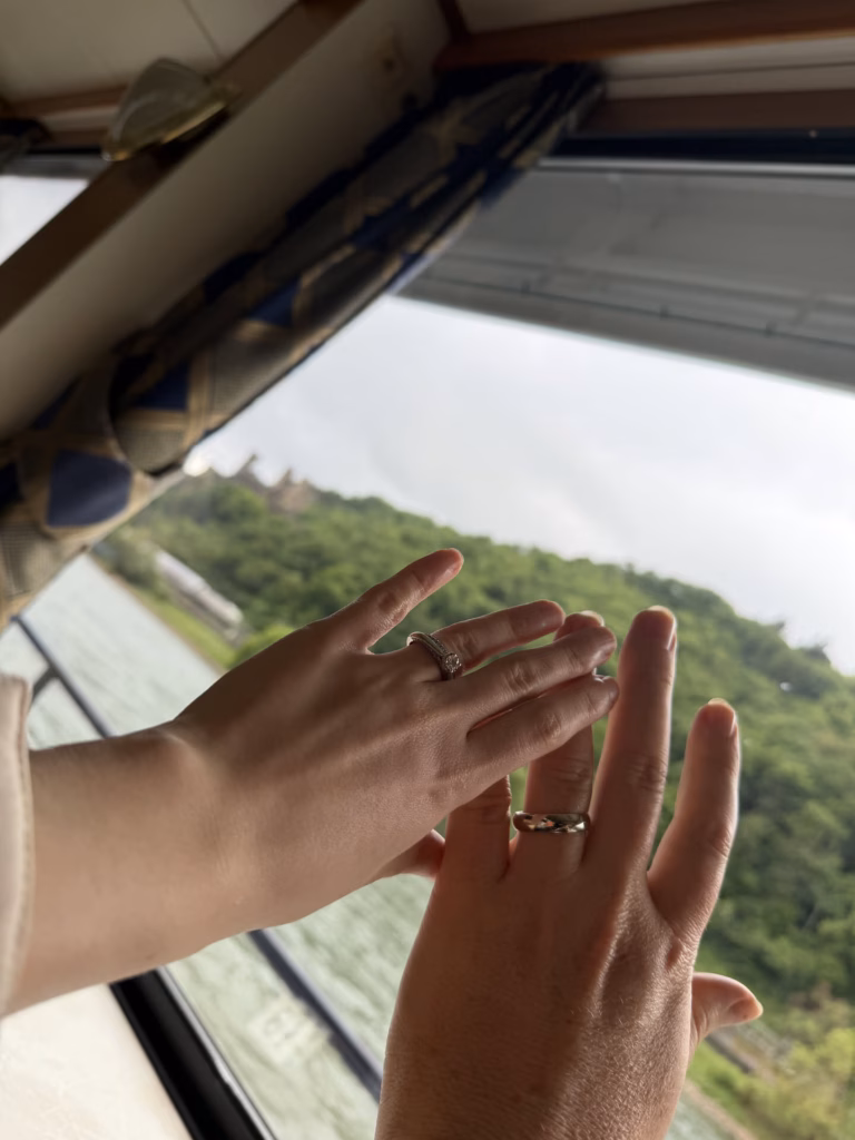 Two hands raised to a boat window, each wearing a wedding ring, green hills and a hilltop castle through the glass