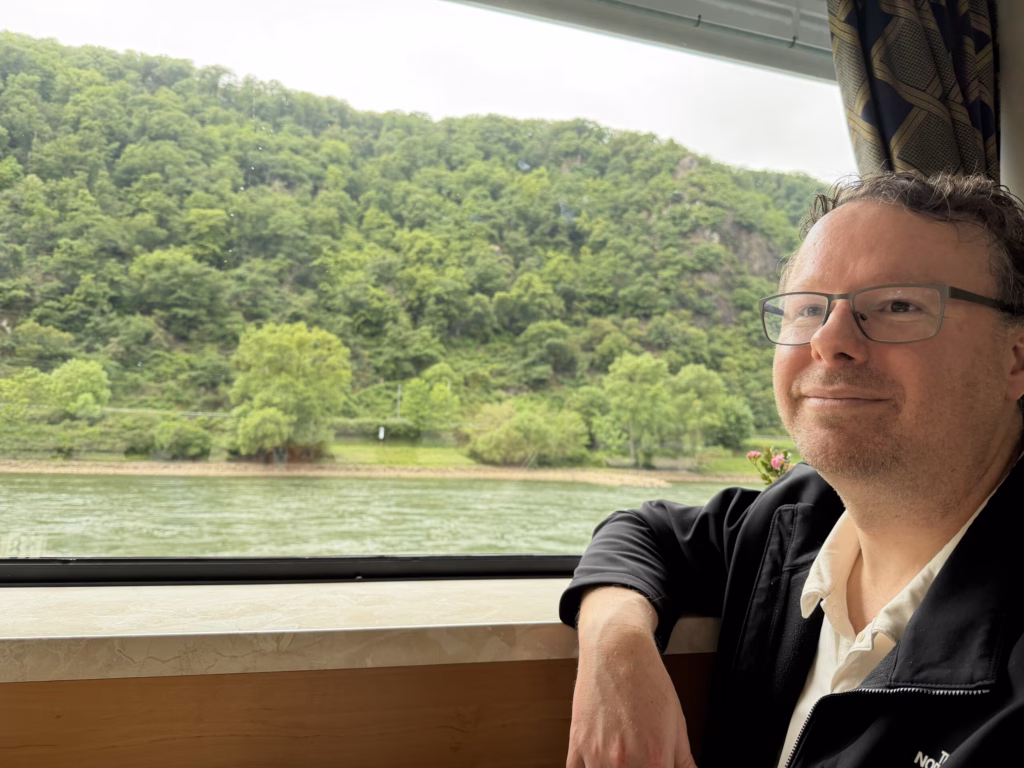 Ryan resting his arm on the window ledge and smiling, the forested gorge wall and river visible through the rain-streaked window