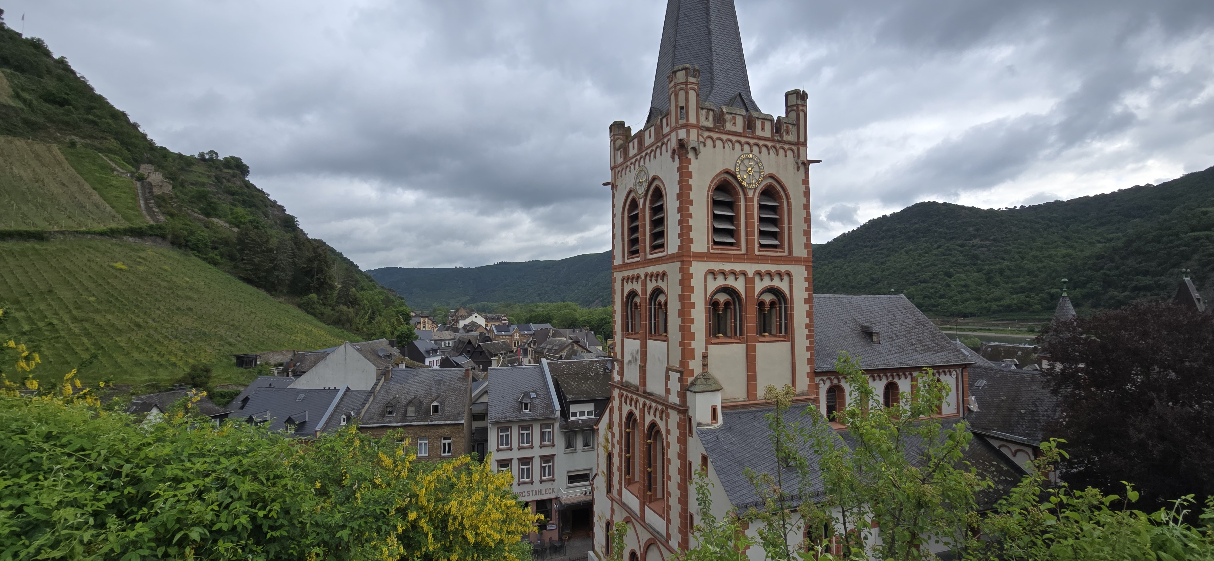 St. Peter Kirche tower from the town wall terrace above the Bacharach Altstadt, Rhine valley hills behind, overcast May sky