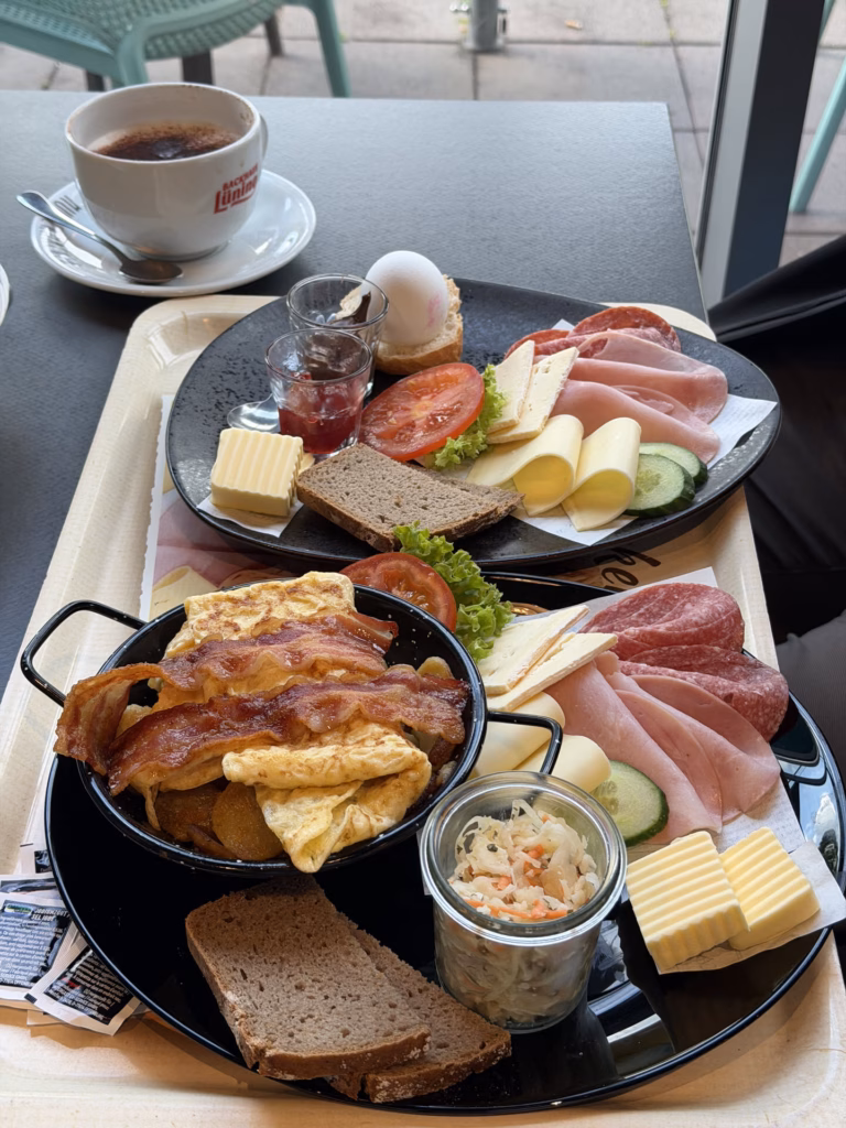 Overhead view of a German breakfast spread with two plates of cold cuts, cheese, tomato and egg, an omelette-and-bacon skillet and coffee