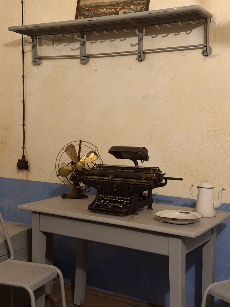 An officer's grey table with a vintage Continental typewriter, a brass desk fan and a white enamel coffee pot and bowl