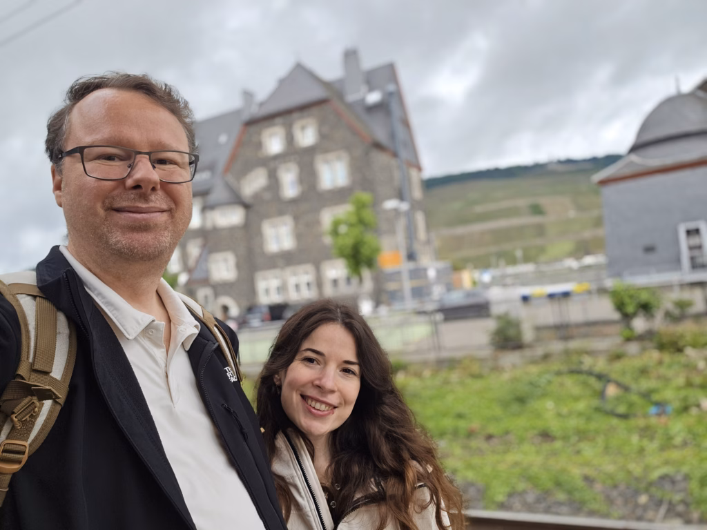 Ryan and Samira taking a selfie at the Bingen Kulturufer railing with the slate pier building, old crane and vineyards behind