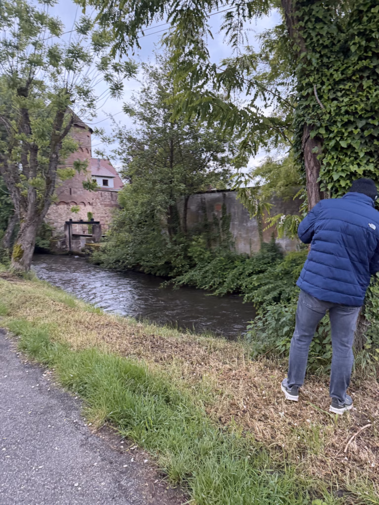Ryan in a navy jacket on the grassy bank photographing the canal toward the round red-sandstone medieval mill tower among willows