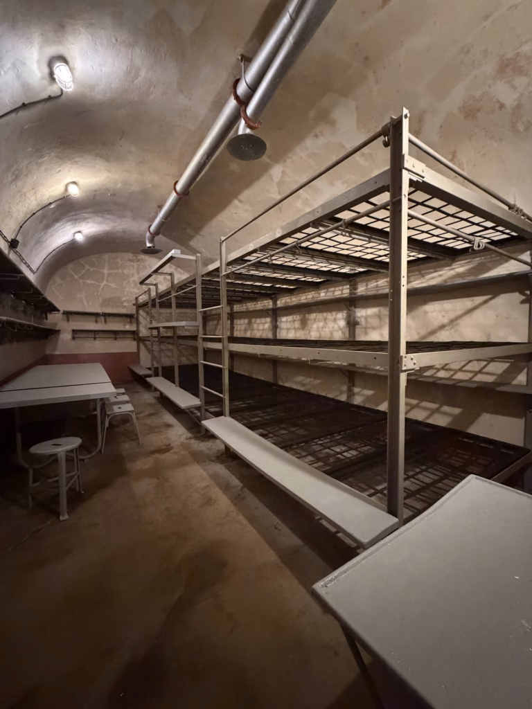 A fort barracks dormitory with rows of three-high metal bunk frames, a table and stools under a vaulted concrete ceiling