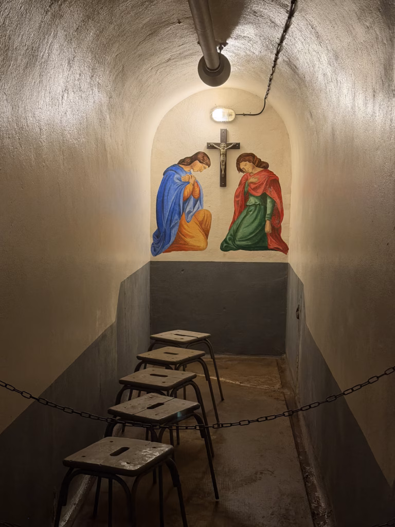 The underground fort chapel with a crucifix and a painted mural of two kneeling praying figures in a vaulted niche, stools below