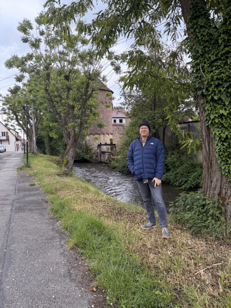Ryan standing on the canal bank with the round red-sandstone mill tower and willows across the green water