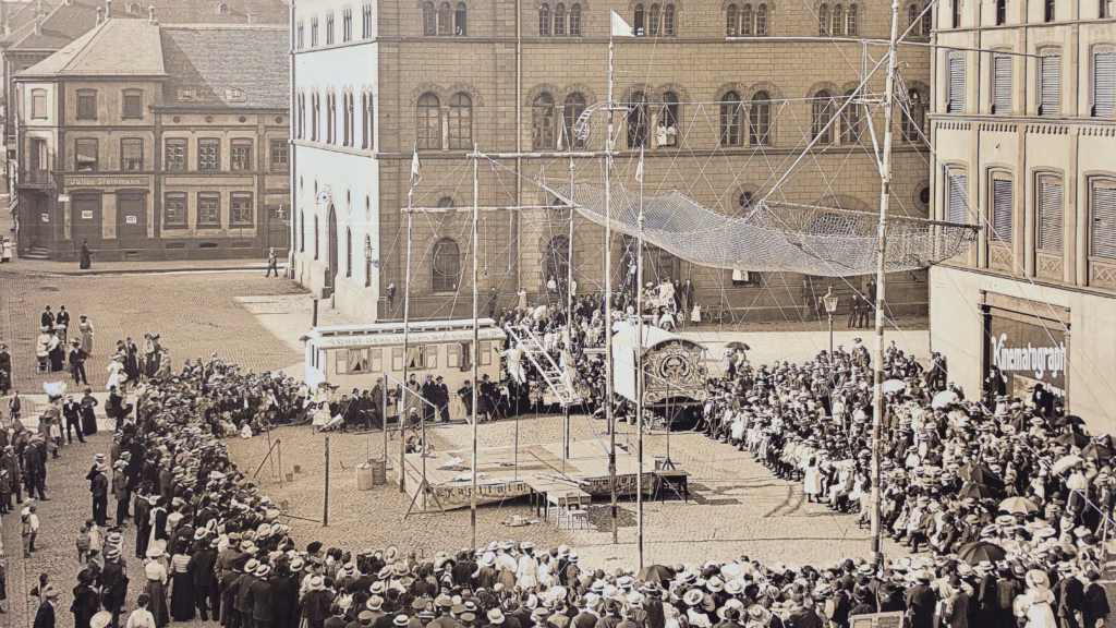 Early-20th-century public event on the Rathaus-Vorplatz, from the Kaiserslautern 750 anniversary display panels: a stage with rigging and a dense crowd in straw hats