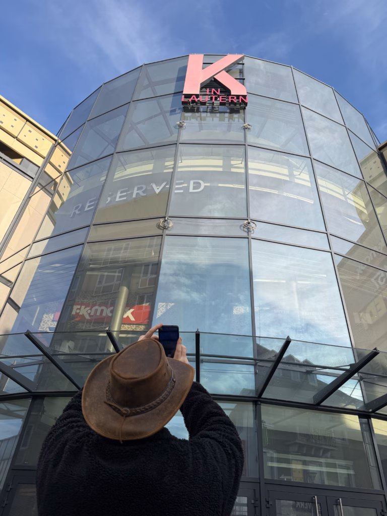 Ryan from behind in leather hat and black fleece, raising his phone to photograph the pink K in Lautern logo atop the curved-glass shopping facade in Kaiserslautern Altstadt, Sunday morning May 2026