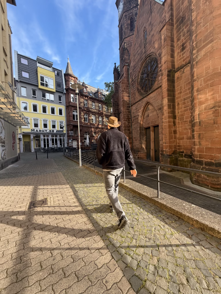 Ryan in leather hat and dark fleece walks down a quiet cobbled alley in Kaiserslautern Altstadt, the red-sandstone east wall of the Stiftskirche rising on his right, yellow Markt 28 cafe townhouse on the left, low Sunday morning light raking the stones