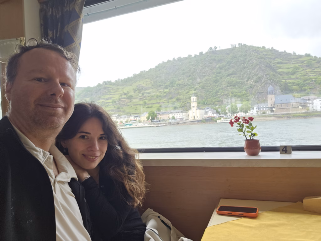 Selfie of Ryan and Samira inside the saloon of the KD southbound cruise boat at Table 4, large window behind them with the table number 4 visible at the right edge and a small red campion flower in a clay pot, St. Goarshausen and the Stiftskirche St. Goar tower visible across the Rhine through the window, gorge cliffs in the background