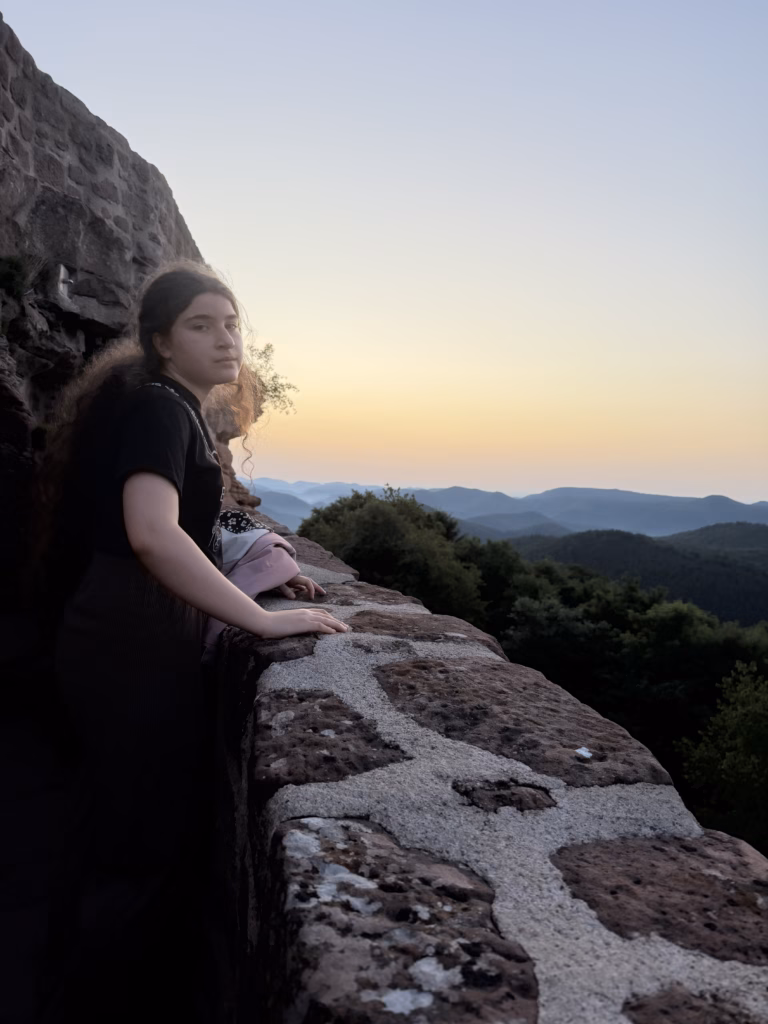 Young woman with long curly hair leaning on the sandstone castle wall at Wegelnburg, looking out over hazy Pfälzerwald hills in pre-dawn light