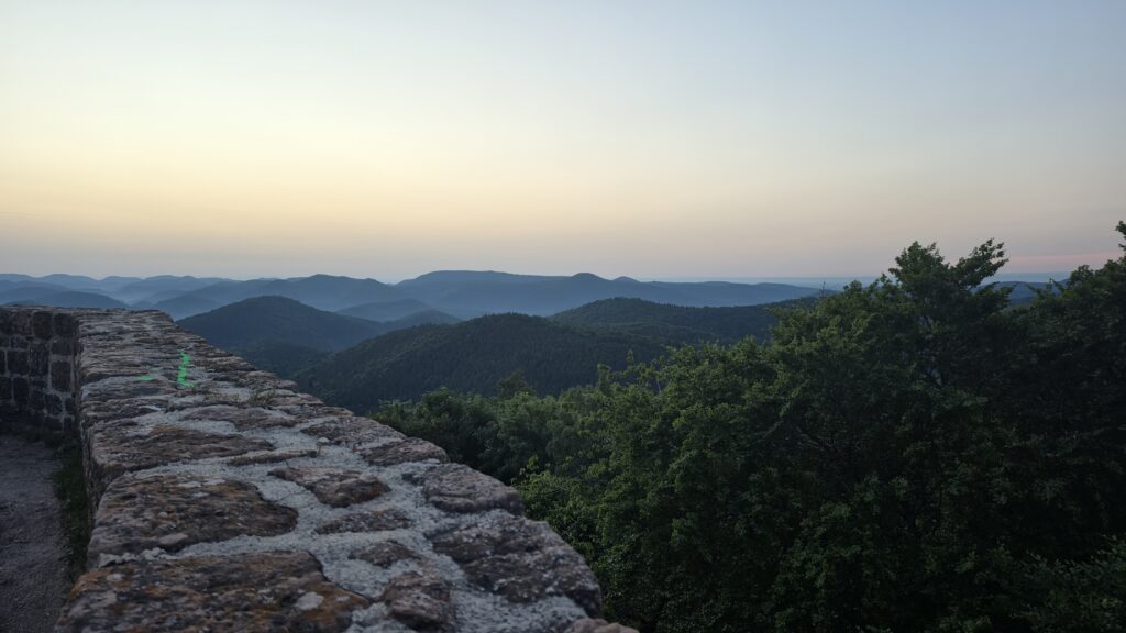 Wide view across the Wasgau mountains from the Wegelnburg ramparts at blue hour, sandstone wall in the foreground, distant horizon glow