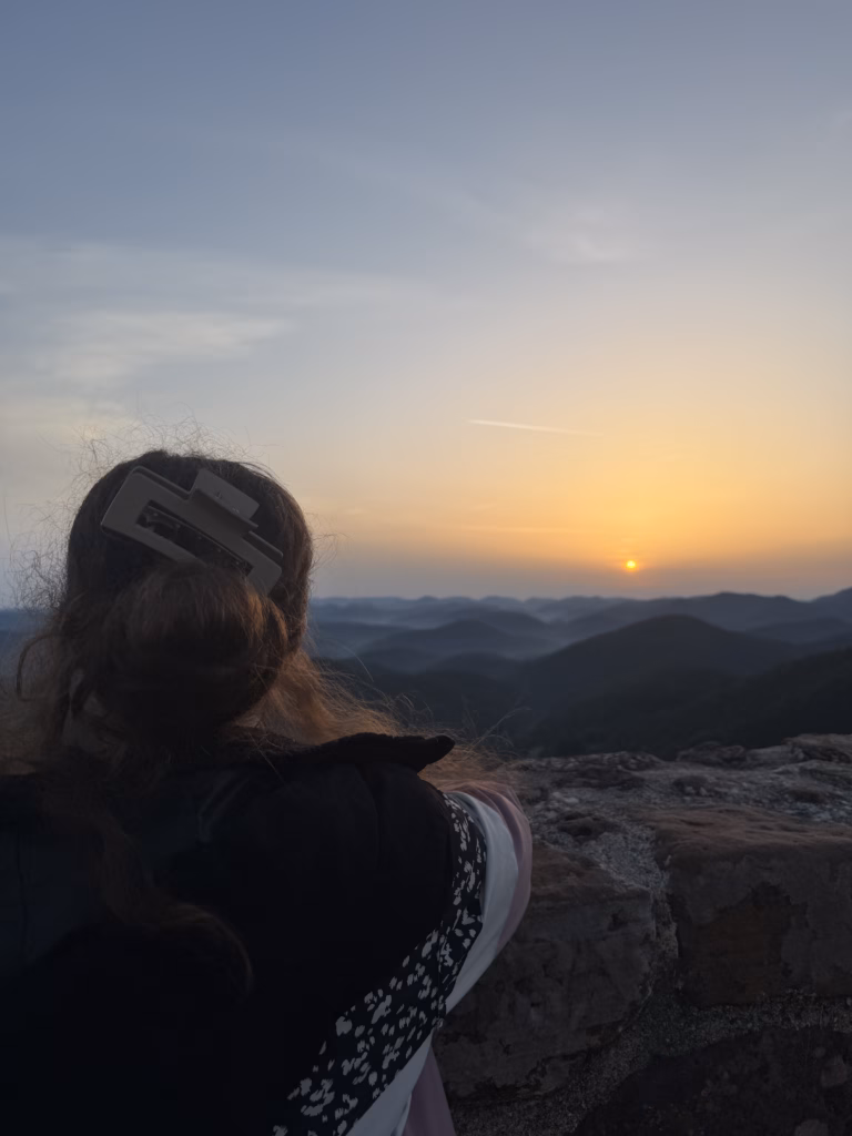 Back of a young woman's head watching the sun emerge over the Wasgau forest from Wegelnburg, pink and gold dawn sky