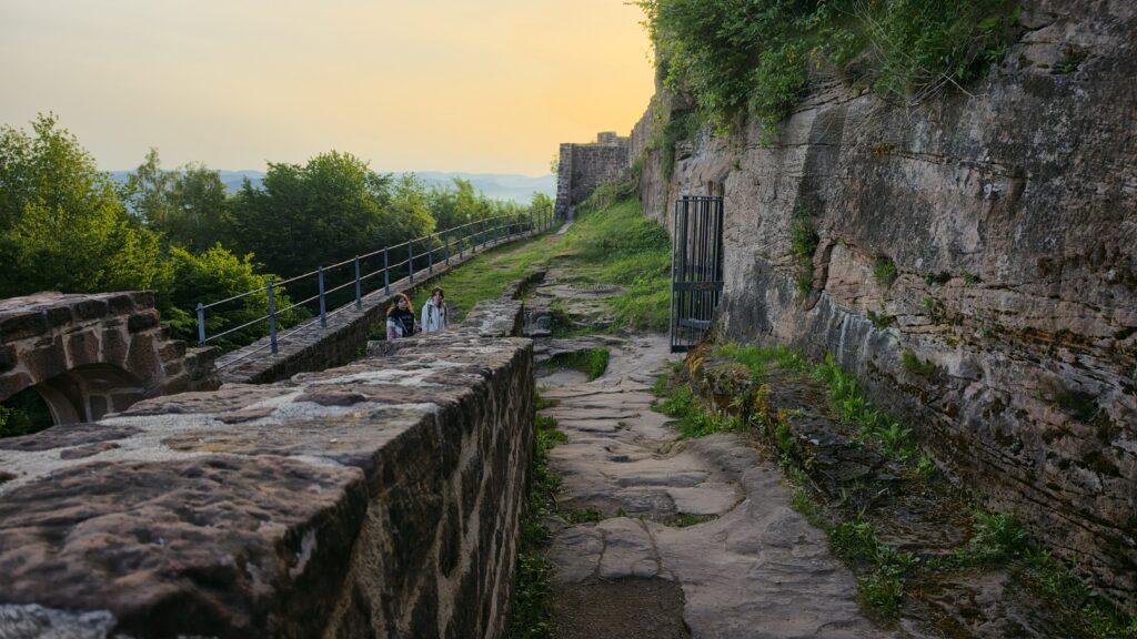 Castle rampart walkway at Wegelnburg with the renovated iron handrail running along the original sandstone wall, two figures seated on the parapet, dawn sky behind