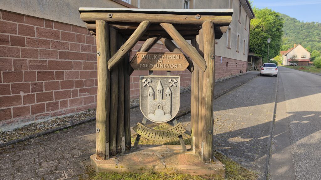 Wooden post-and-beam village welcome shelter at the Ortseingang of Nothweiler bearing the village crest with castle iconography and the sign Willkommen im Erholungsort Nothweiler