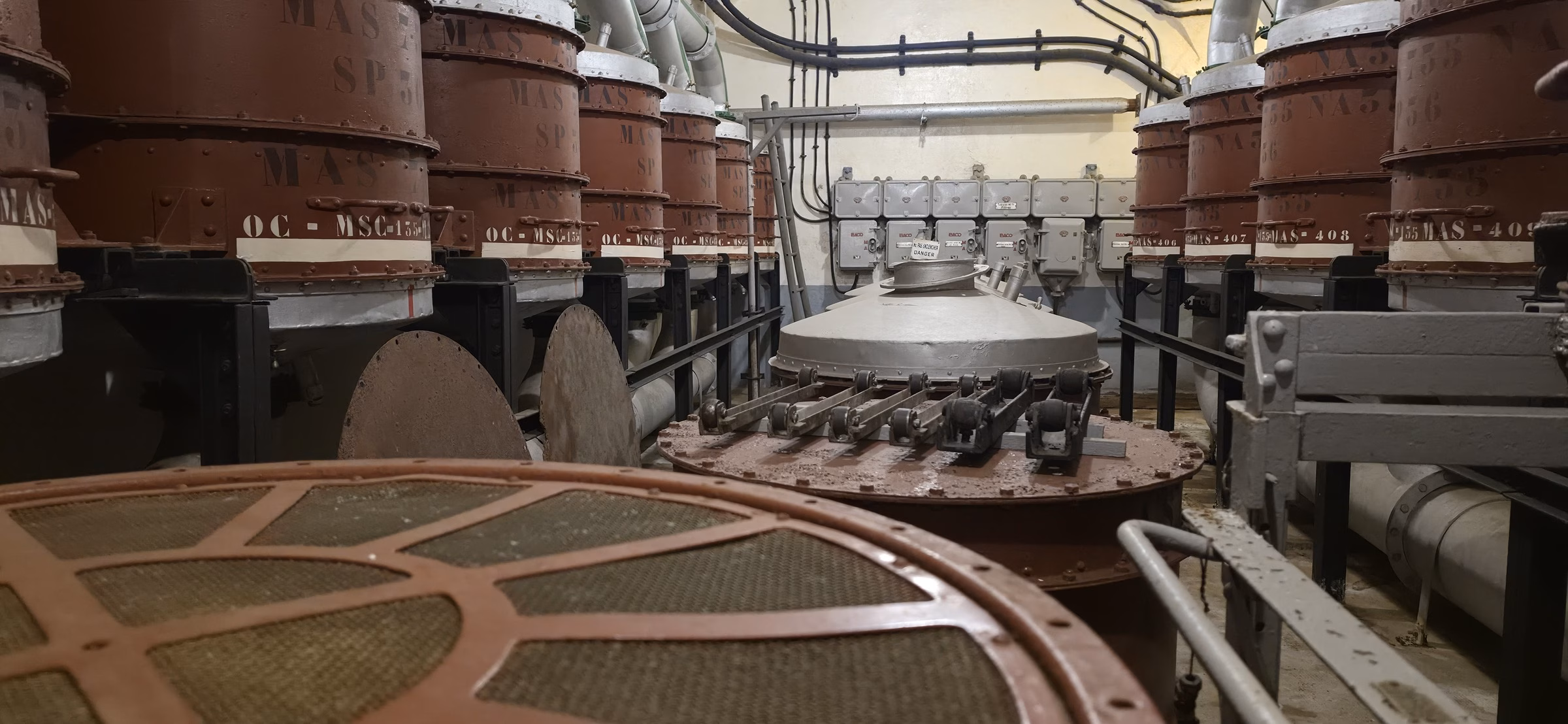Large vertical carbon-and-filter drums lining the back wall of the salle des filtres at Fort Schoenenbourg, with circular fan housings set into the concrete floor in the foreground
