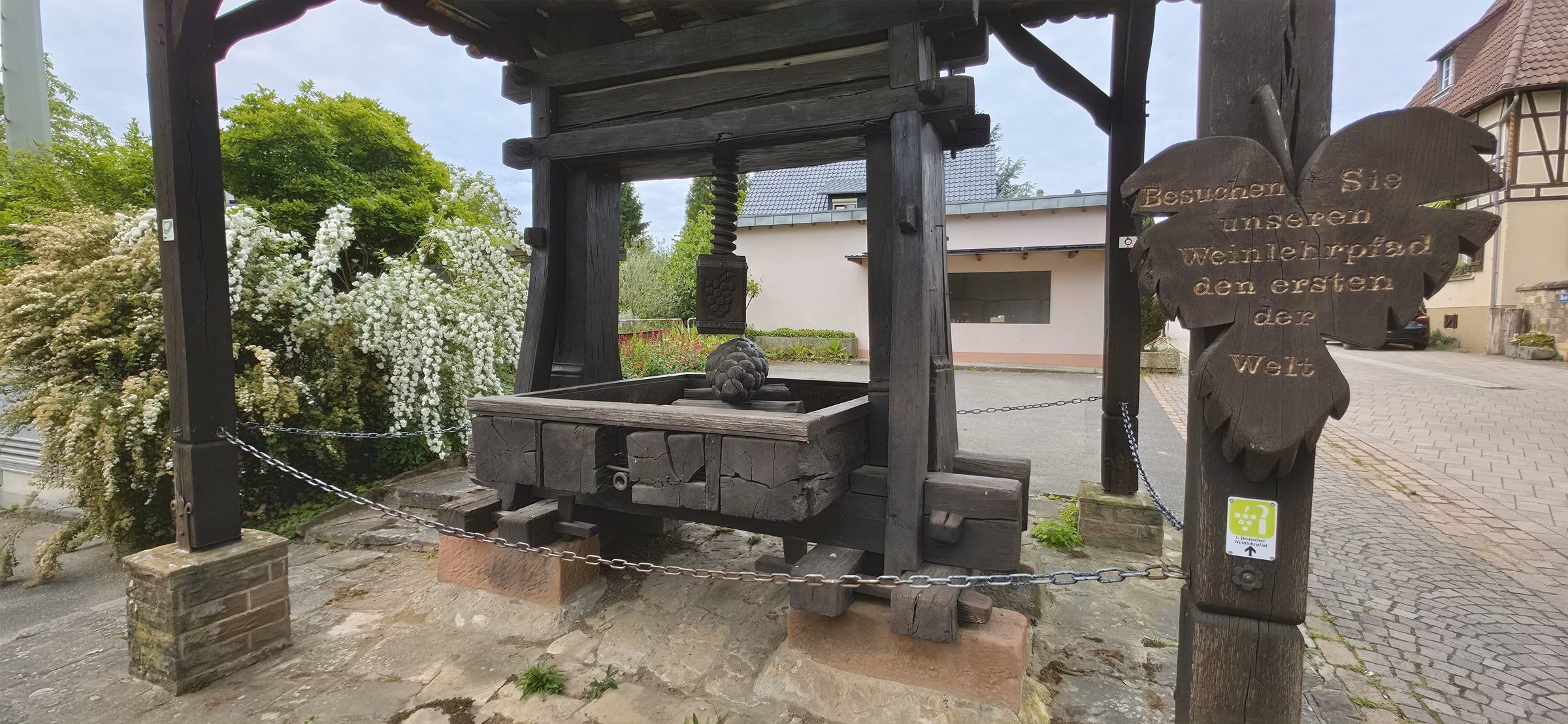 Historic wooden wine press at the Deutsches Weintor courtyard with a wooden Weinlehrpfad sign marking the start of Germany's first educational wine trail